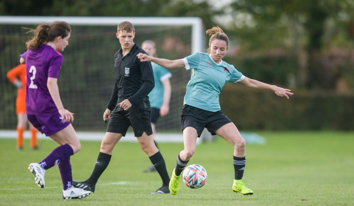 Eponine in a light blue shirt and black shorts is in control of the ball during a match. She is being closely watched by a young male referee in black and two opposing players in purple jerseys. The background is a well-maintained grassy soccer field.