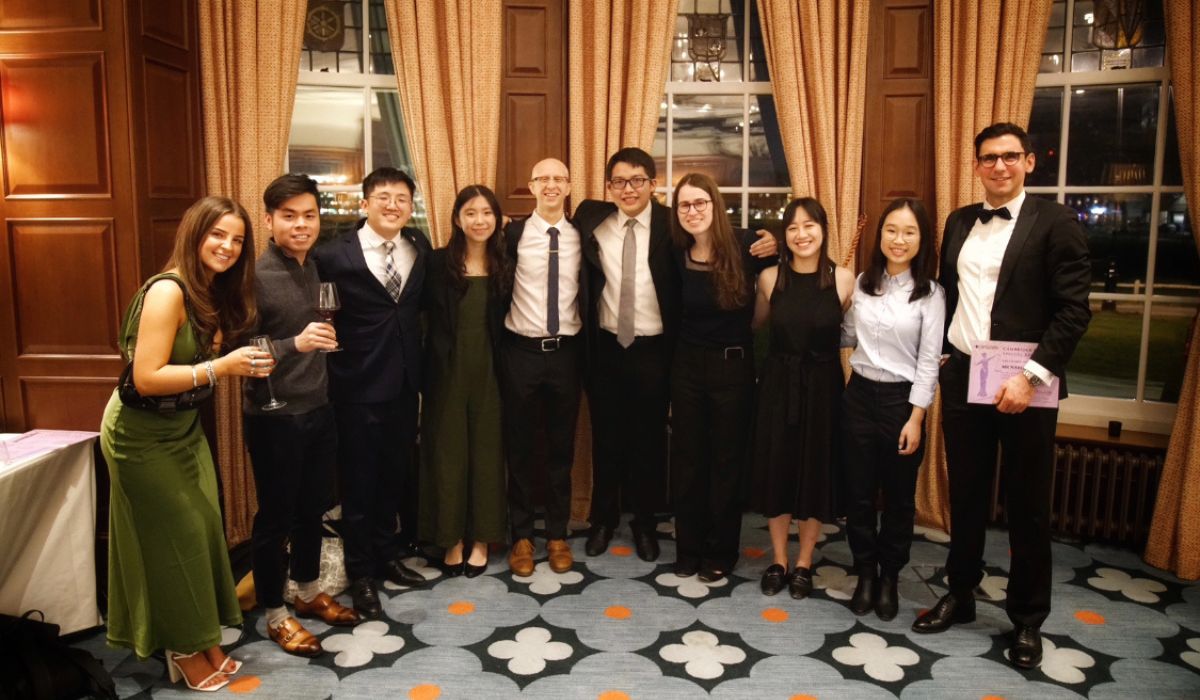 Nabil and ten students, standing in a room on a patterned carpet, with curtains and a large window in the background