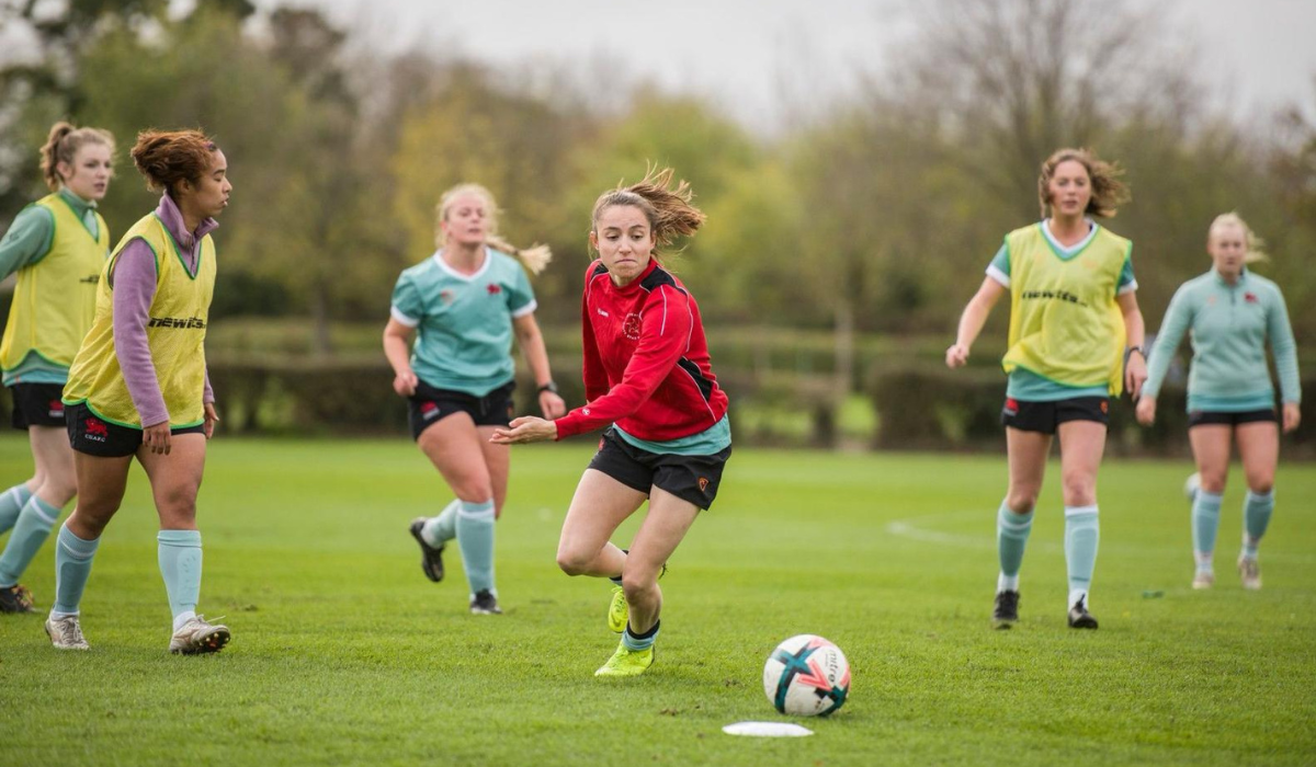 Eponine in a red and black uniform kicking a football during a practice session on a grassy field, with teammates in various colored vests