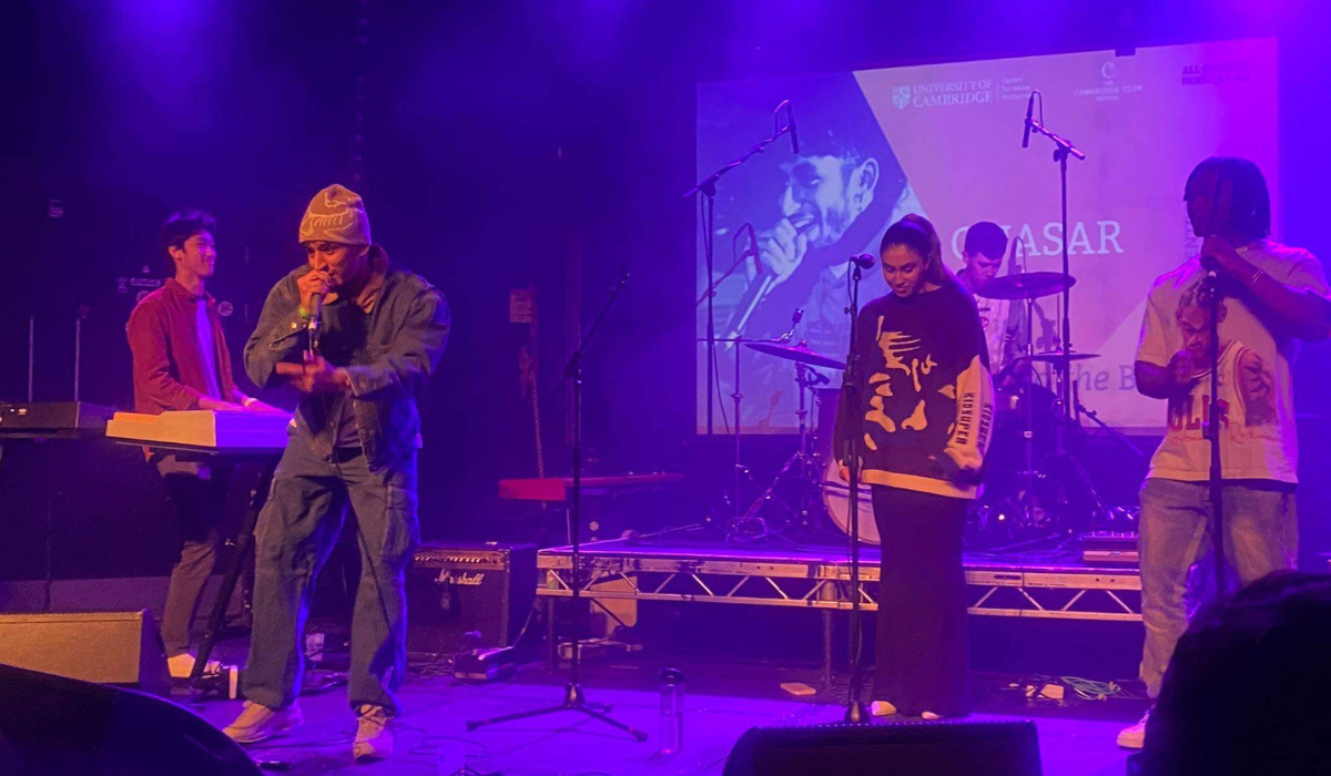 The image features a band performing on stage. There is a large screen in the background displaying a visual, possibly relating to the event or the band itself. There are five visible members: a keyboardist, a vocalist, a guitarist, a drummer, and another vocalist or speaker. They are illuminated by stage lighting, which gives the scene a vibrant atmosphere typical of live music events. There is a University of Cambridge logo.