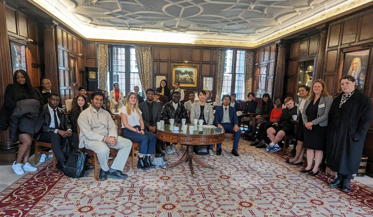 A diverse group of young adults, dressed in formal attire, seated and standing in an ornate wood-paneled room with a decorative ceiling and a red patterned carpet. They are gathered around a circular table with mugs. The room has large windows with heavy drapes, framed paintings on the walls, and a portrait visible on the right. Everyone is looking towards the camera, smiling.