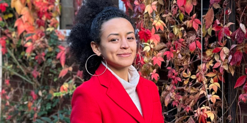 Maria Morena posing for a photo in front of a wall covered in red leaves, wearing a red coat and hair tied up in a ponytail