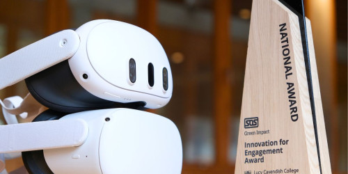 The image shows two white virtual reality headsets one on top of the other resting on a surface. Adjacent to the right, there's a wooden triangular award plaque with text that reads "NATIONAL AWARD", below which is a logo and additional text that appears to say "Innovation for Engagement Award" followed by "Lucy Cavendish College University of Cambridge". 