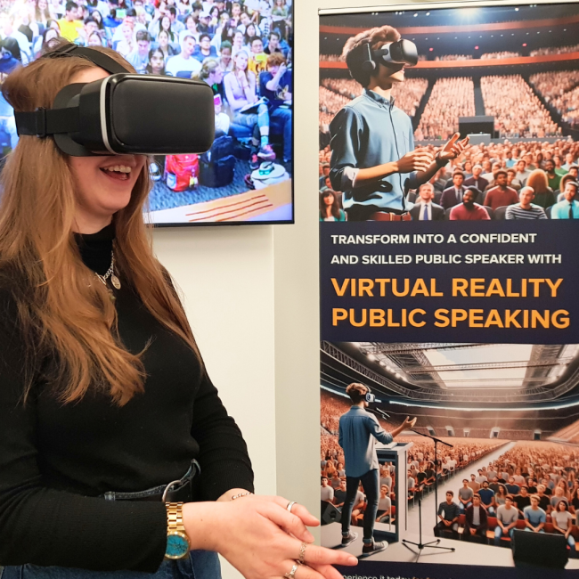 A woman wearing a virtual reality headset smiles while immersed in a VR experience. Behind her is a large screen showing an image of a crowded auditorium, and next to her is a banner promoting "Virtual Reality Public Speaking." The banner features an illustration of a person wearing a VR headset, practicing public speaking in front of a large virtual audience, with the tagline: "Transform into a confident and skilled public speaker with Virtual Reality Public Speaking.